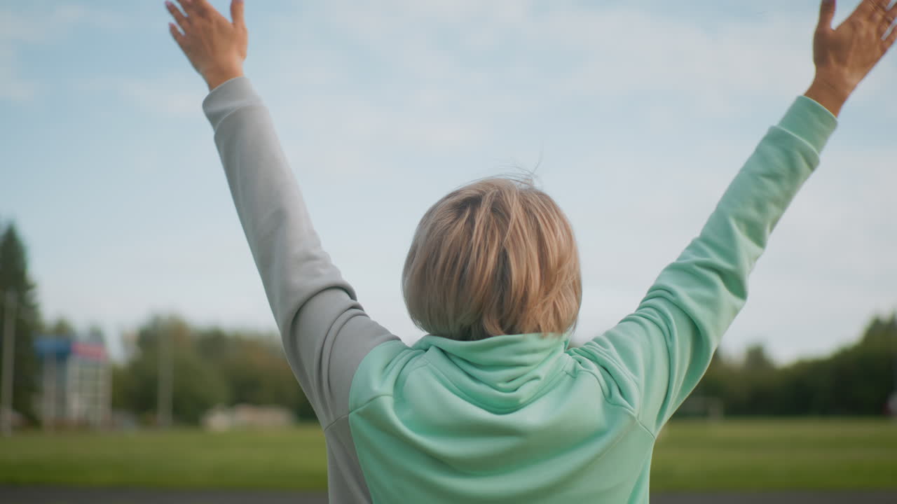 Back view of sports woman wearing ash hooded sweatshirt standing in green garden arms open eyes closed practicing deep breath work embracing peaceful outdoor moment under bright sky with lush trees