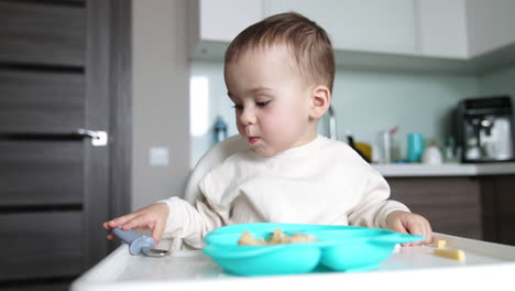 mealtime for a lovely cute caucasian baby. kid sits at feeding table with plate in front of him. child takes a spoon.