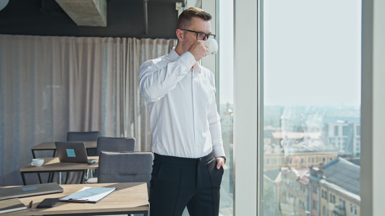 Pensive confident male entrepreneur drinking coffee standing at the window in the office. Businessman having lunch enjoying city view.