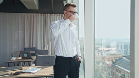 pensive confident male entrepreneur drinking coffee standing at the window in the office. businessman having lunch enjoying city view.