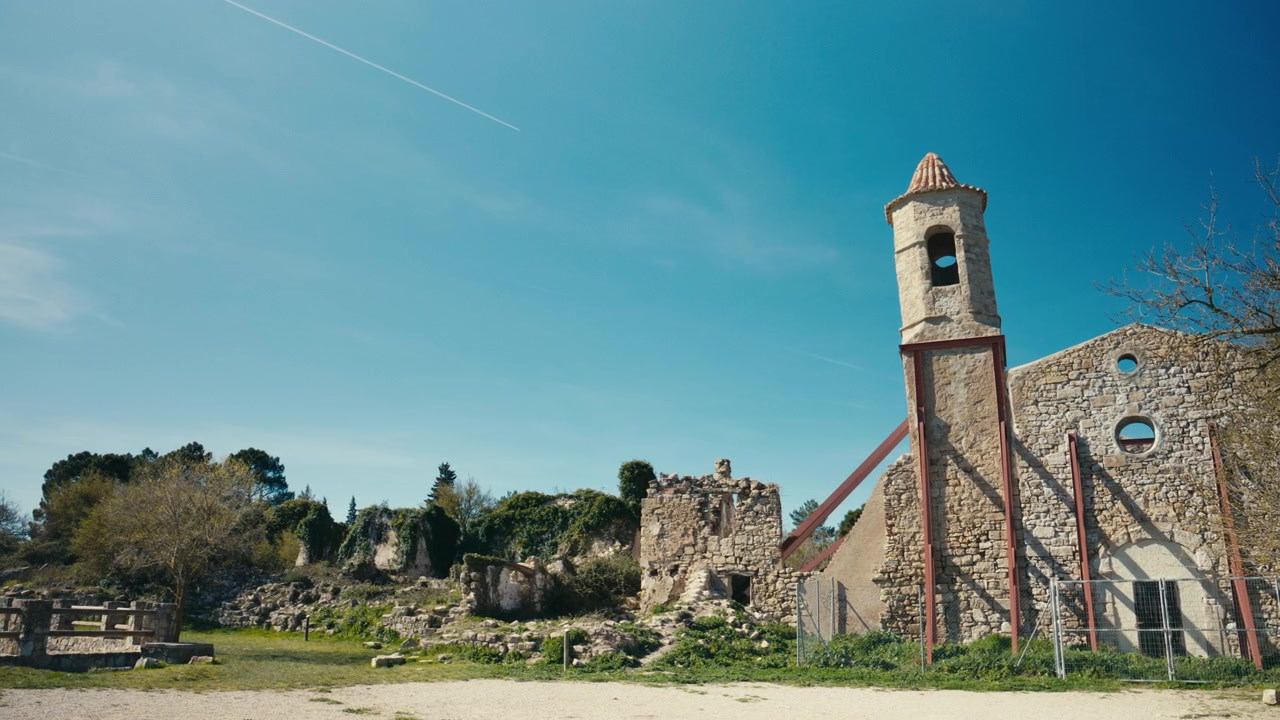 Un campanile in pietra consumato dal tempo si erge accanto ai resti di una vecchia chiesa, creando una suggestiva scena storica con muri in rovina, terreno aperto e cielo azzurro brillante.