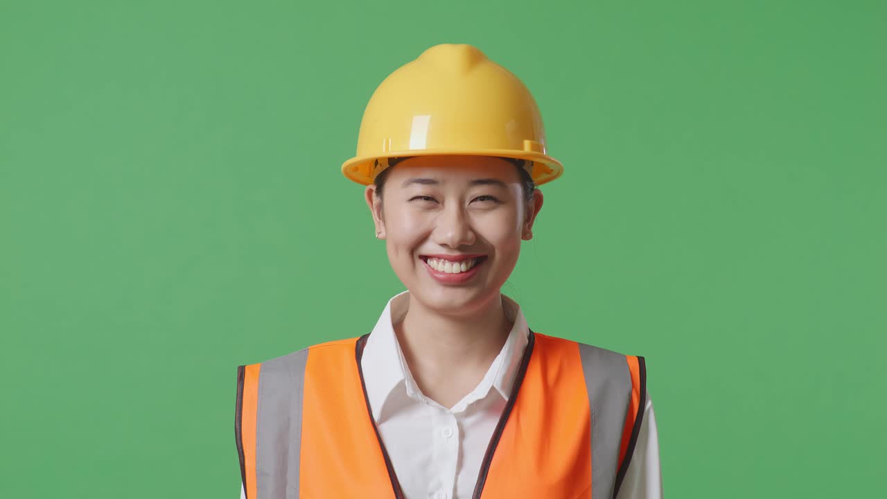 안전 헬을 입은 아시아 여성 엔지니어가 녹색 스크린 배경 스튜디오에 서서 카메라를 향해 미소 짓고 있습니다.