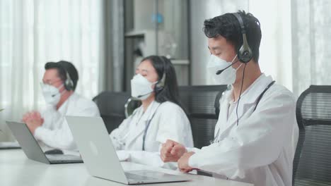 three asian doctors with stethoscopes in headset and masks working as call center agents sitting on chairs and being happy due to success working with computers at the office