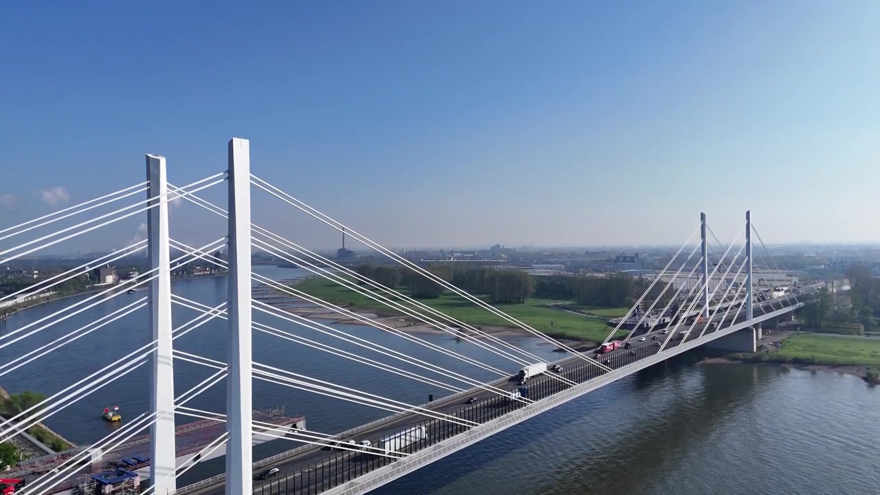 Rising aerial perspective over the Rhine, showing the diagonal bridge, cars, trucks, river, and distant industrial smoke.