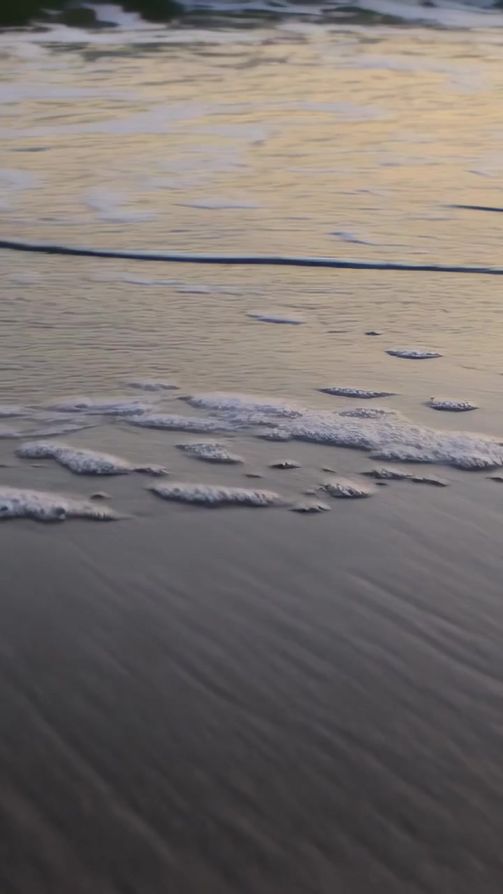 วิดีโอแนวตั้ง: คลื่นเล็กๆ ซัดเข้ามาพัดฟองสีขาวไปตามหาดทรายเปียกในช่วงพระอาทิตย์ขึ้น