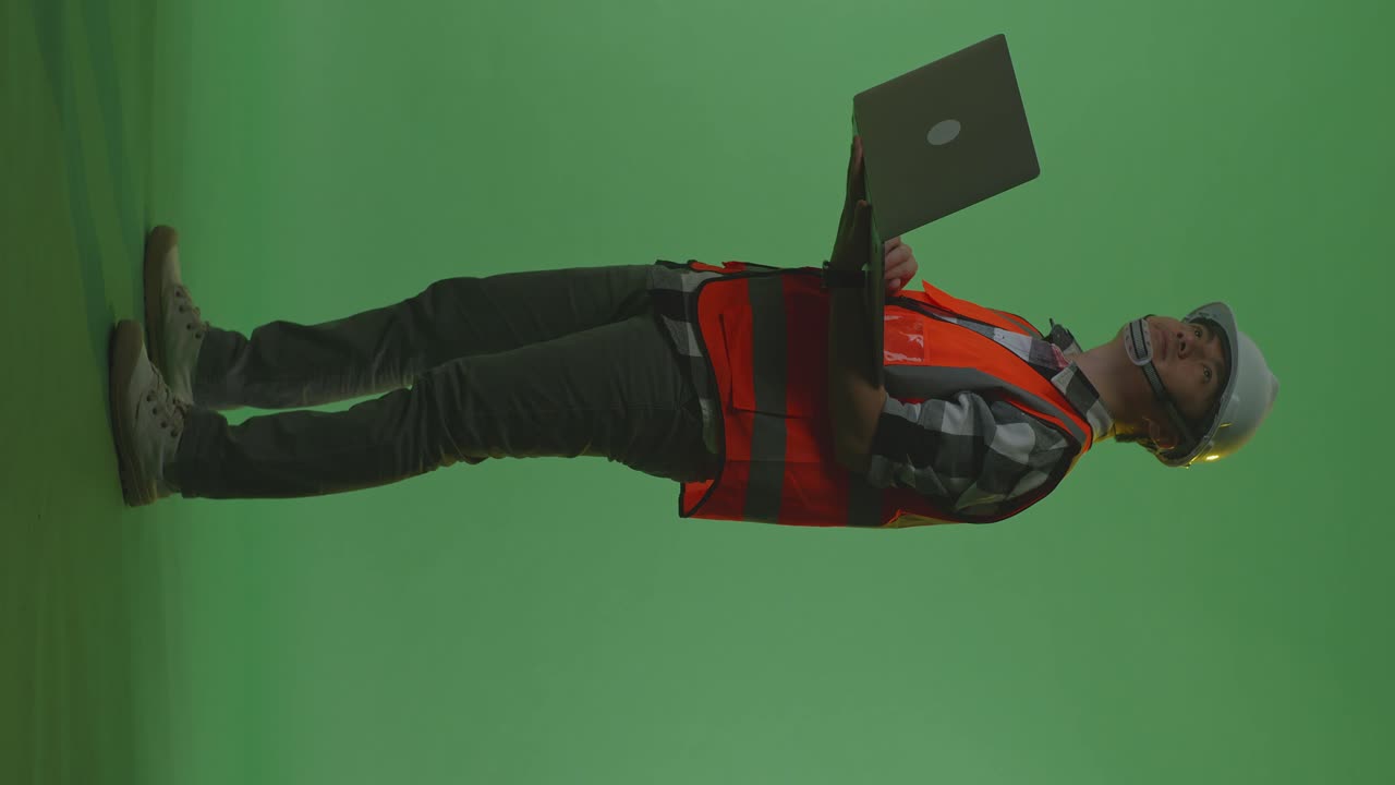 vista lateral completa del cuerpo de un ingeniero masculino asiático con casco de seguridad trabajando en una computadora portátil y mirando a su alrededor mientras está de pie en el estudio de fondo de pantalla verde, durante la puesta o el amanecer