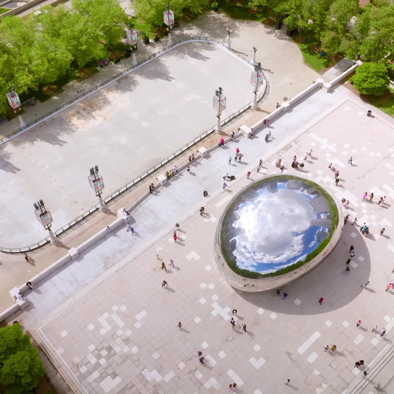People walking around famous Cloud Gate sculpture in Chicago. The Bean reflecting sky, clouds, park and skyscrapers in its top surface. Top view