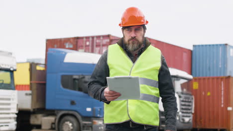 lavoratore che indossa giubbotto e casco di sicurezza che organizza una flotta di camion in un parco logistico mentre consulta un documento e cammina