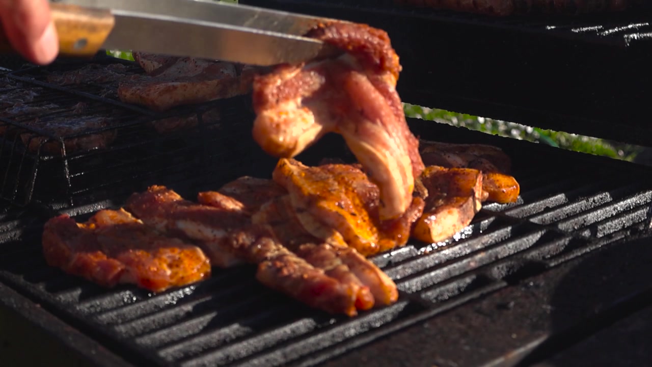Deliziose costine di maiale condite che cuociono su una griglia calda per barbecue al rallentatore, con una persona che usa pinze e un coltello per sistemare la carne sfrigolante mentre un denso fumo sale dalla carbonella