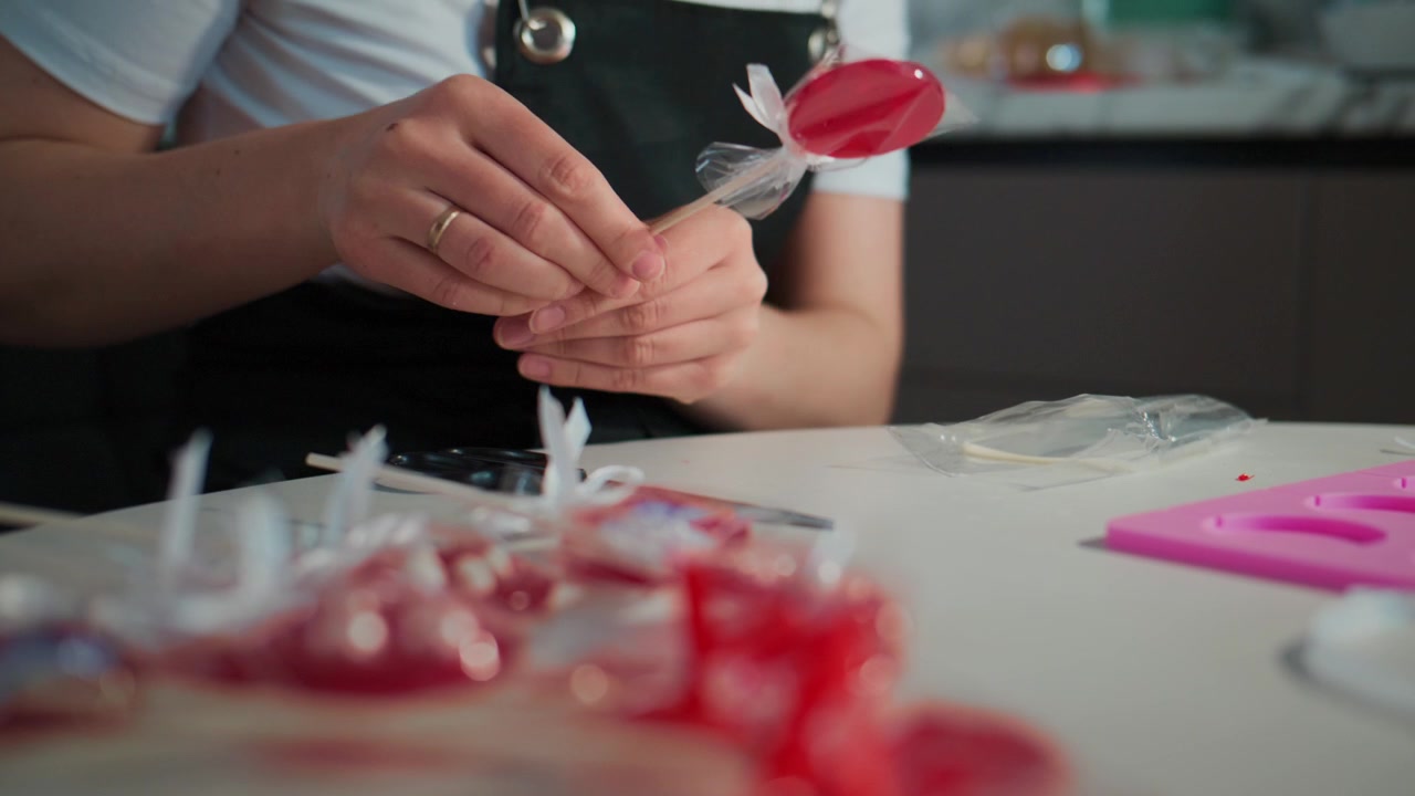 Person in an apron carefully cuts red candy wrappers with scissors. The workspace is cluttered with colorful candy pieces. The individual focuses intently on precise cutting