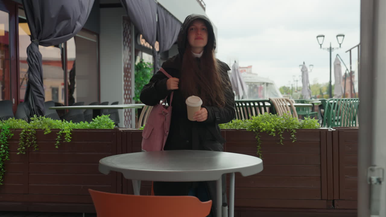 white woman walking away from cafe hooded coat and pink backpack carrying takeaway cup, empty terrace table and chairs, city skyline under overcast sky, slow reflective pace of student commuter