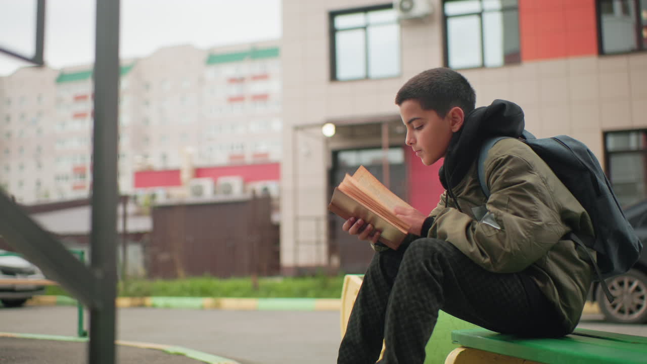 Side view of young kid seated with backpack studying book outdoors on bench with blurred urban building in background and glowing bulb near door post showing focus