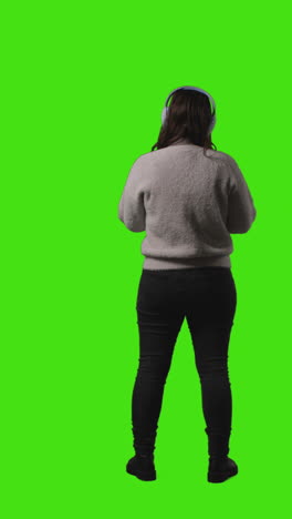 vertical video rear view full length shot of woman wearing wireless headphones streaming music from mobile phone against green screen 1