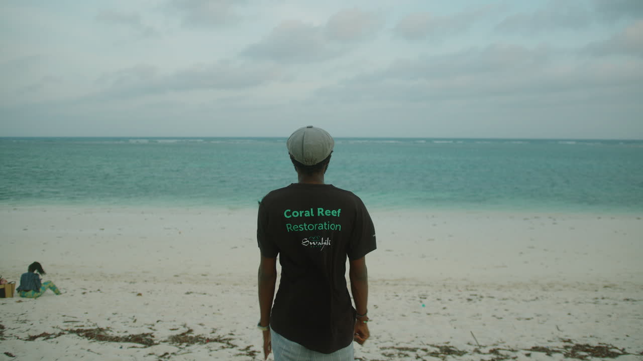 General medium shot of a young black man on the shore of a beach with his back to the camera and looking out to sea