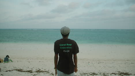 general medium shot of a young black man on the shore of a beach with his back to the camera and looking out to sea