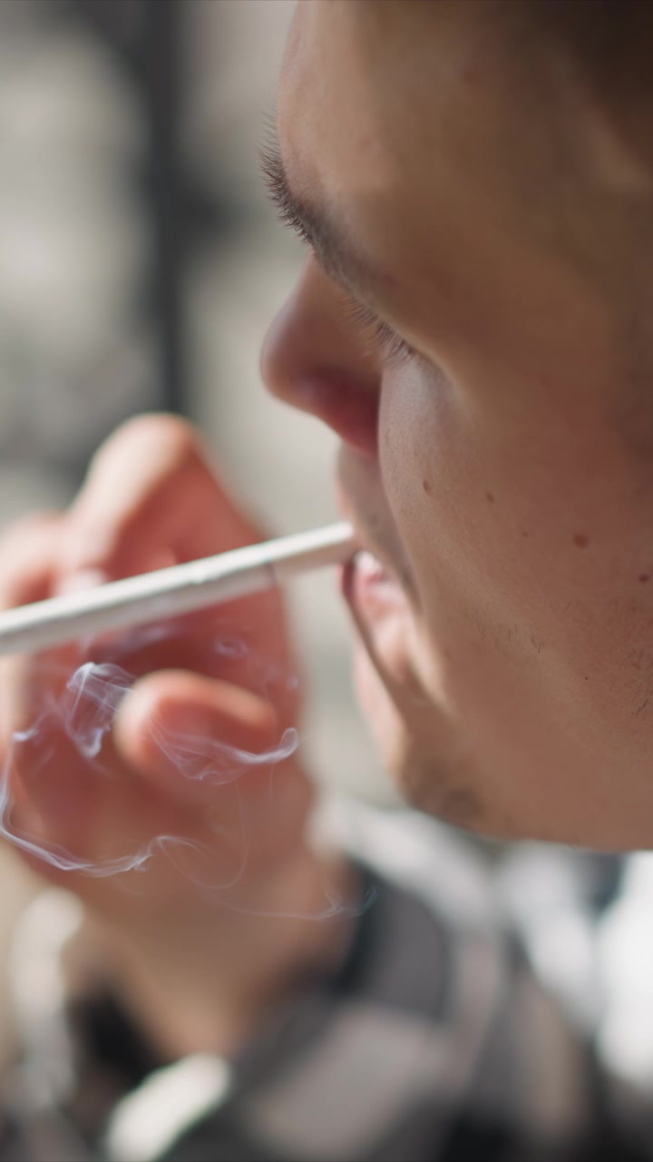 Primo piano di un uomo che espira fumo da una sigaretta elettronica. Il soggetto tiene il dispositivo vicino alla bocca con lineamenti del viso visibili e un leggero movimento mentre inala il vapore. L'illuminazione soffusa crea