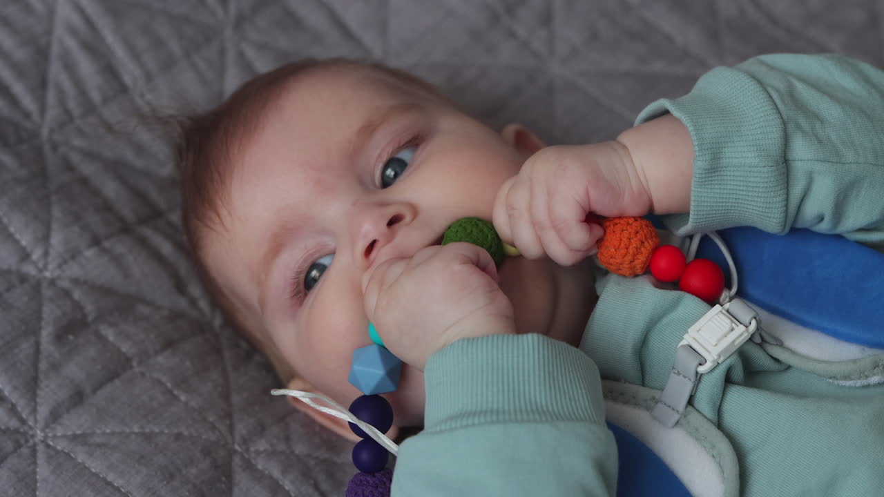 Young baby girl lying on back, chewing on chew toy of colorful shapes
