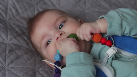 young baby girl lying on back, chewing on chew toy of colorful shapes