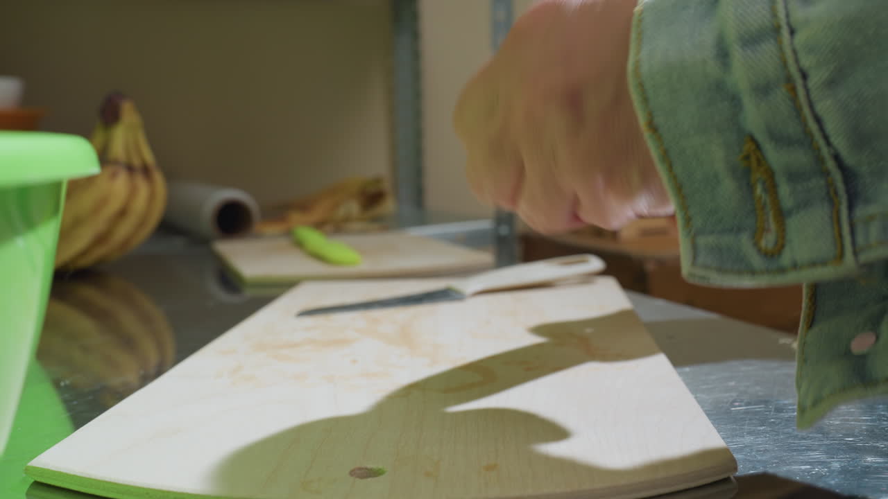 houten plank, gepelde banaan op aanrecht met groene kom op de achtergrond, handen die fruit schillen met een vaste beweging, provisiekast en tros bananen zichtbaar, intieme close-up die de textuur laat zien