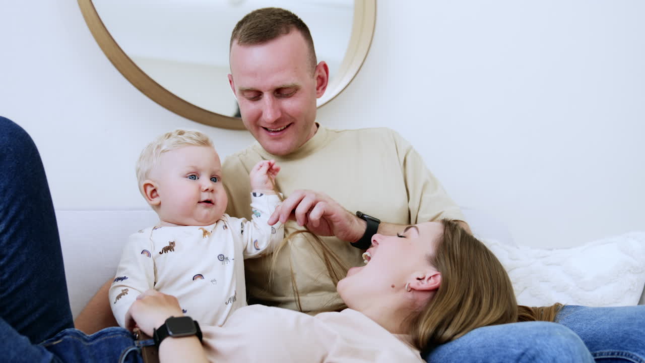 Lovely young family of three are all on the sofa at home. Woman lies on man's laps and baby boy touches mom's hair. Low angle view.