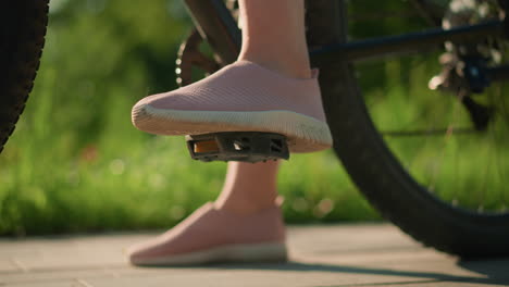 close-up of leg in pink sneakers gently tapping bicycle pedal, while other foot rests on ground, cyclist prepares for ride, blurred background shows park greenery