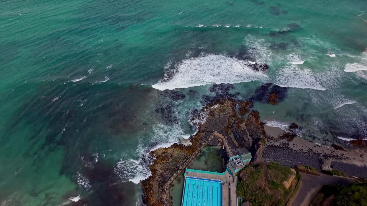 Drone top-down shot moving toward the ocean, showing a saltwater swimming pool by the coast, waves breaking on rocks, and a nearby road at St Kilda Beach in Dunedin