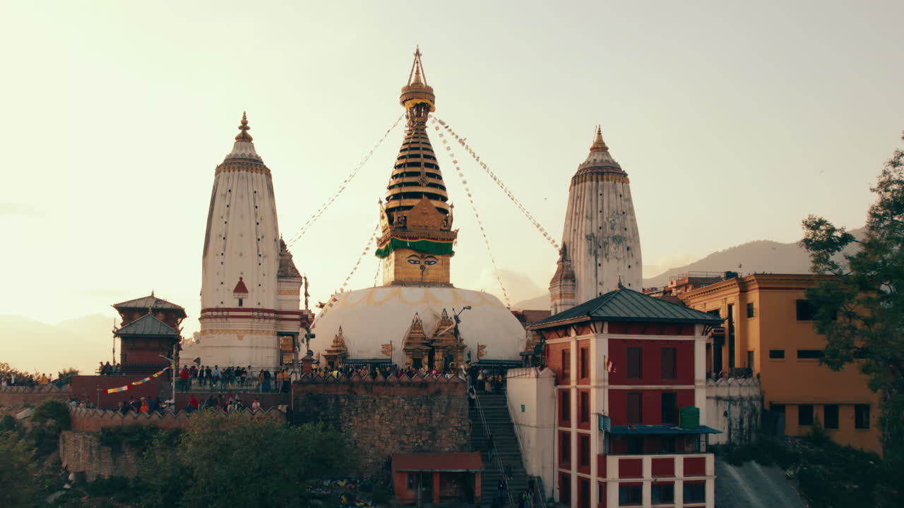 Starting close on the iconic Swayambhunath Stupa, the drone shot smoothly pans out, revealing the stunning panoramic view of the Monkey Temple with its vibrant golden hues basking in the evening sky.