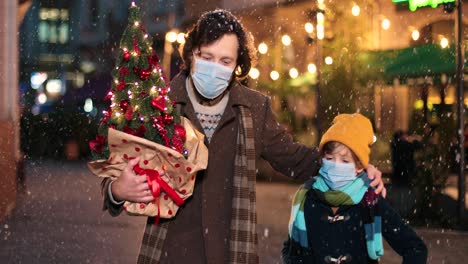 close-up view of caucasian happy father and son wearing facial masks walking down the street and holding christmas tree and present while it¬¥s snowing in christmas