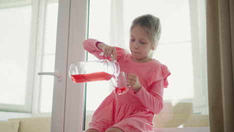 young girl wearing pink dress sitting by window pouring red juice from glass jar into clear mug spilling drink on dress indoors with soft blur background