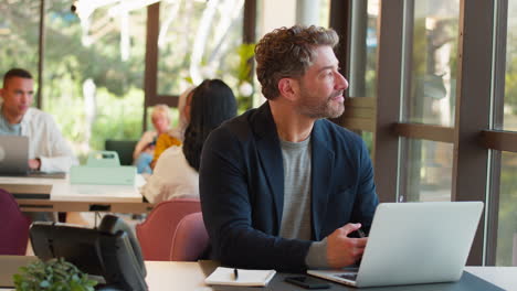 Mature-Businessman-Wearing-Glasses-Working-In-Office-Writing-In-Notebook-And-Looking-Out-Of-Window