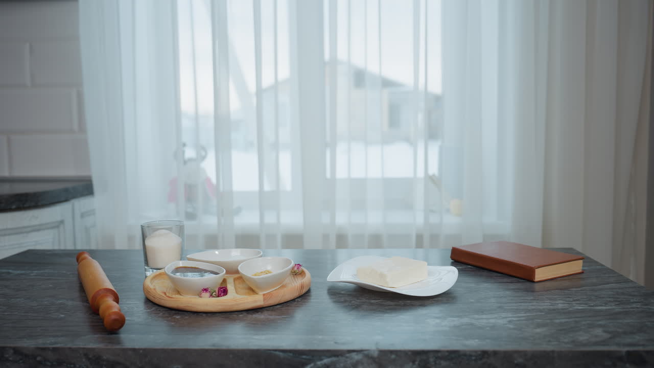 우아한 검은색 주방 테이블, 나무 트레이, 접시, 향신료, 주방 도구, 갈색 커버 책, 접시 위에 버터 에 놓여, 색 한 커튼, 창문으로 건물 밖을 드러낸다.