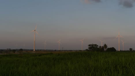 Wind-Turbines-shooting-out-of-a-Farmland-while-the-Sun-is-setting-as-Trucks-Pass-by-making-Clouds-of-Dust,-clean-alternative-energy-in-Thailand-and-mainland-Southeast-Asia