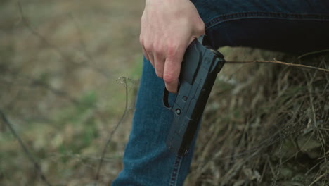 a close view of a person holding a black handgun, their hand resting on their knee, with a blurred background of dry grass and soil