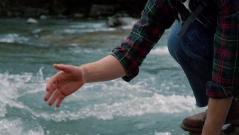 tourist taking break during hike at river. man touching clear water with hand