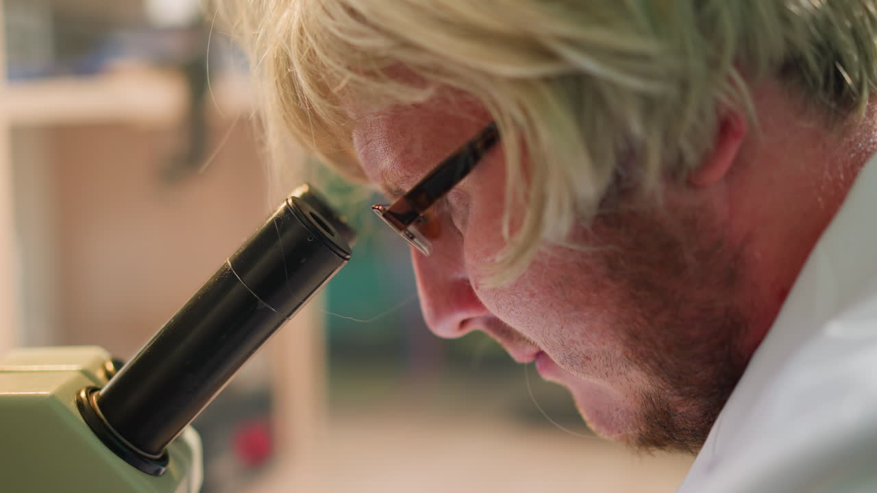 금발 가발과 안경을 입은 과학자가 미세경을 통해 실험실에서 미세한 배경을 가진 표본을 집중적으로 조사하는 근접 시각