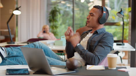 Young-Businessman-With-Feet-On-Desk-In-Office-Listens-To-Music-On-Wireless-Headphones-Air-Drumming