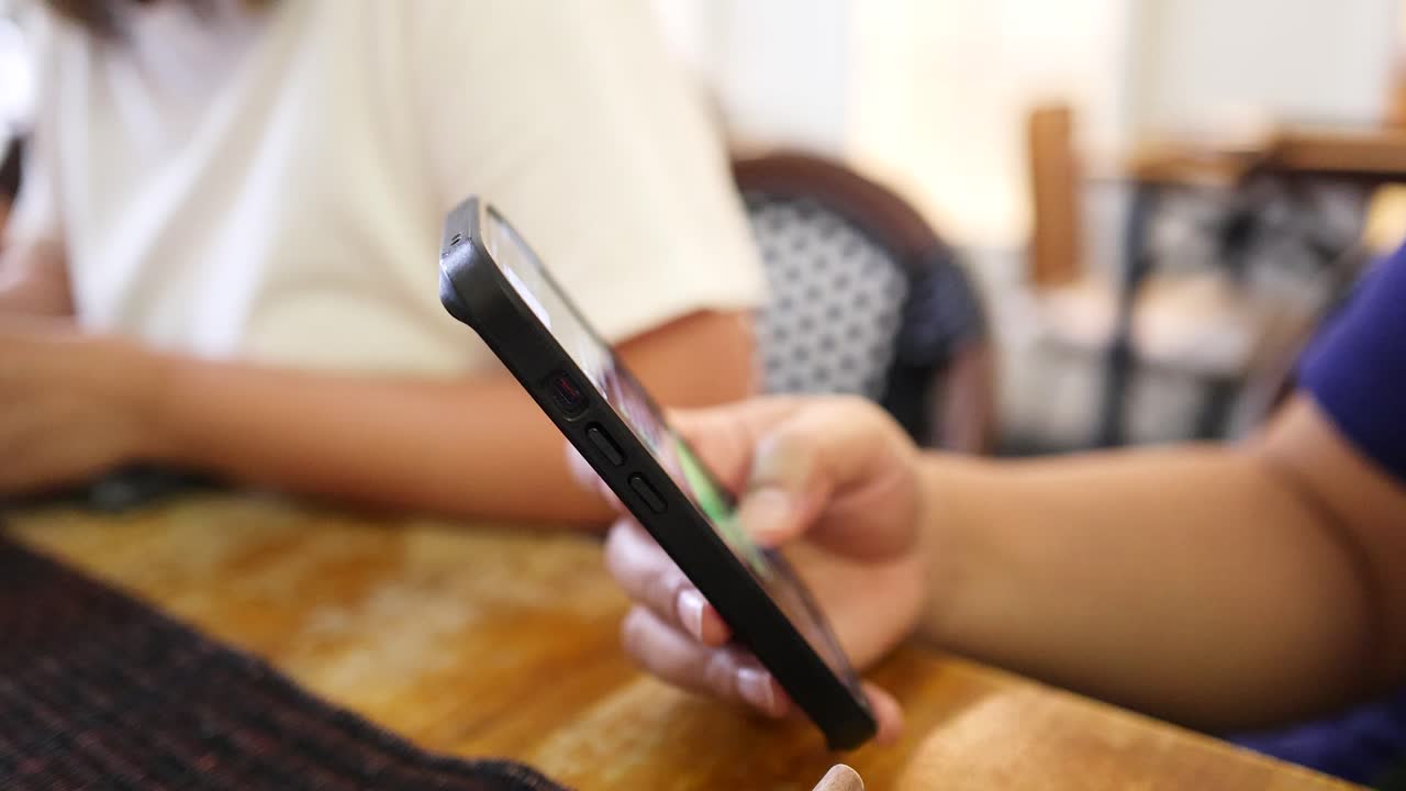 A person interacts with a smartphone using one hand while seated at a wooden table in a softly lit indoor setting, with shallow depth of field