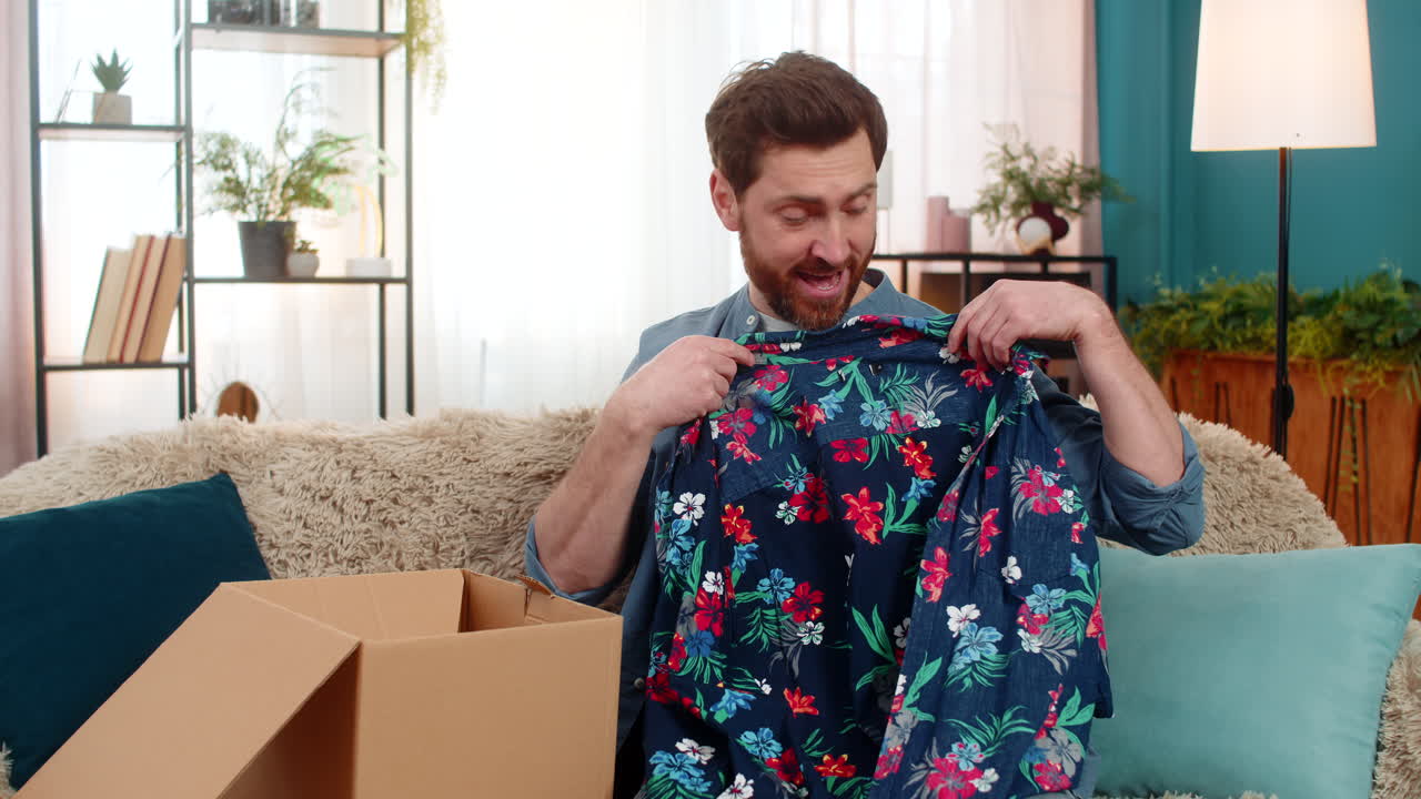 Young man excitedly unpacks a package pulls out a colorful shirt and examines it with delight