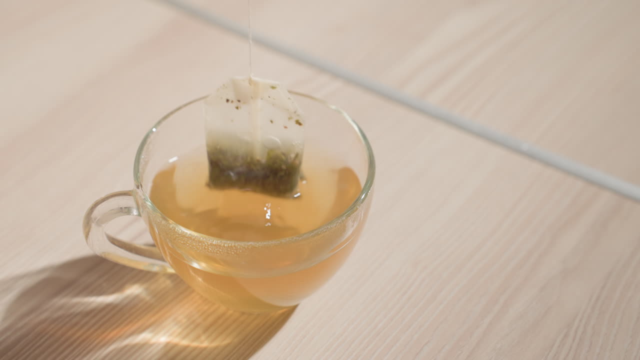 Close-up shot of hand holding tea bag above glass cup with steeping herbal tea, releasing the tea into the cup. Gentle lighting creates calm and relaxing atmosphere in minimal kitchen setting