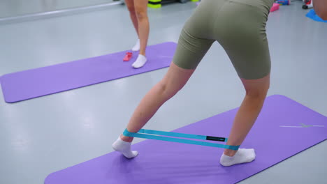 close-up, vista de ángulo bajo de una mujer haciendo ejercicio con bandas de resistencia en una alfombra de yoga púrpura, otra persona de pie cerca, equipos de gimnasia visibles en el fondo borroso