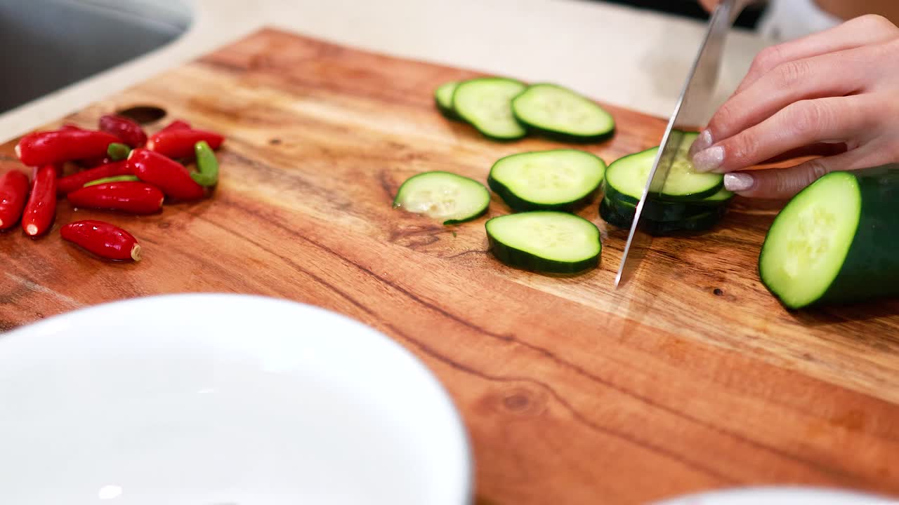 A person slices cucumbers on a wooden board in a bright kitchen, surrounded by fresh vegetables