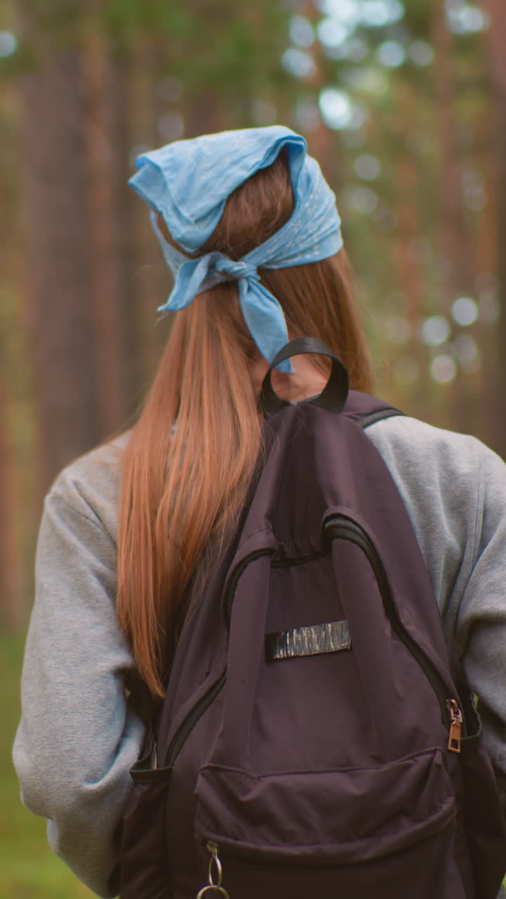 задний вид молодой женщины, проходящей через пышный лес, в рюкзаке и синей бандане, руки, держащие ремни рюкзака, в окружении высоких деревьев и яркой зелени