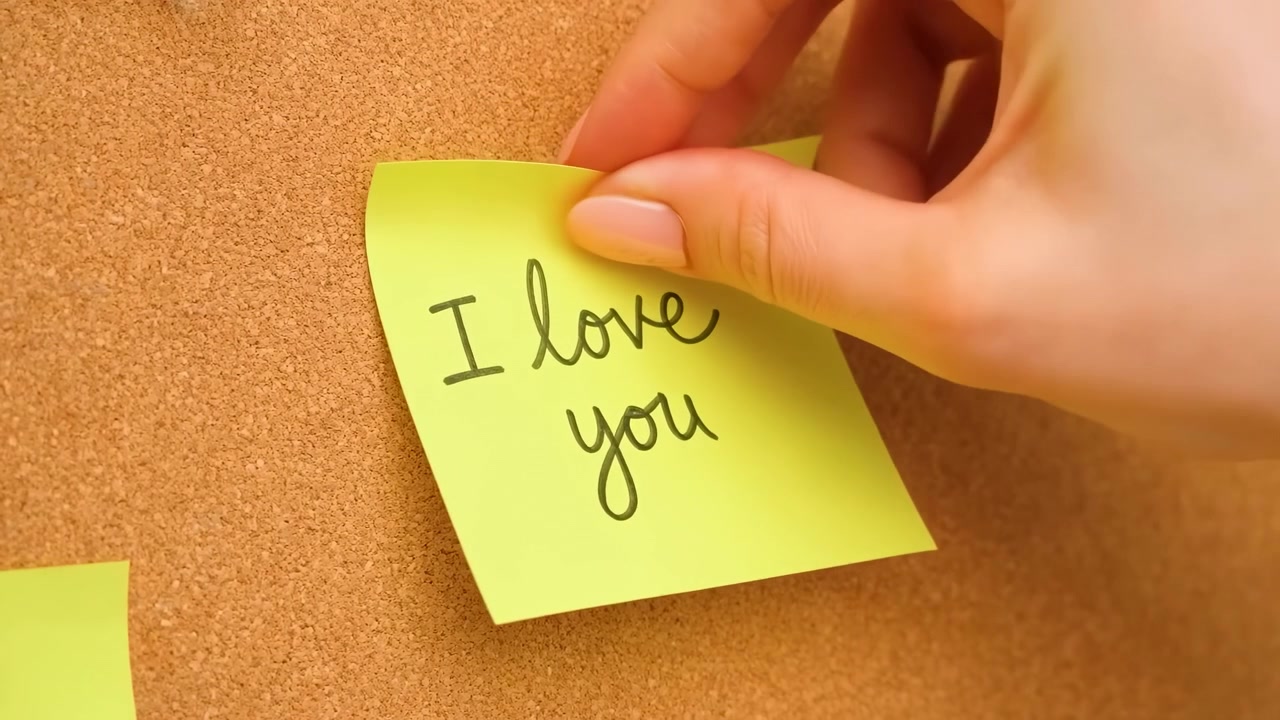 Scena in primo piano in movimento di una mano curata che posiziona un post-it giallo brillante su una bacheca di sughero testurizzata, con contatto visibile delle dita, curvatura della carta, messaggio scritto a mano e un'atmosfera calda da ufficio