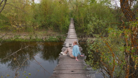 mujer sentada en un viejo puente de madera sobre un río en un bosque