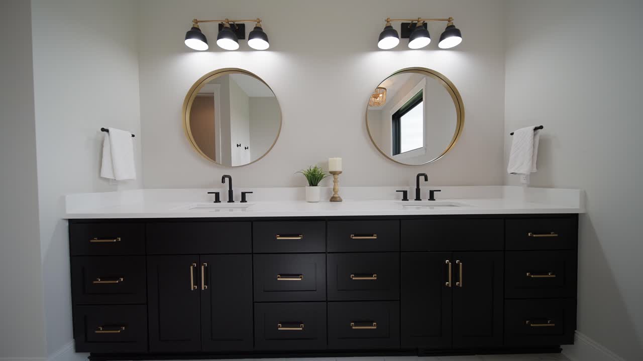 Wide, straight on push-in shot of a white and bright bathroom with white floors, countertops, 2 black faucets, black cupboards, a green plant, and 2 circular mirrors