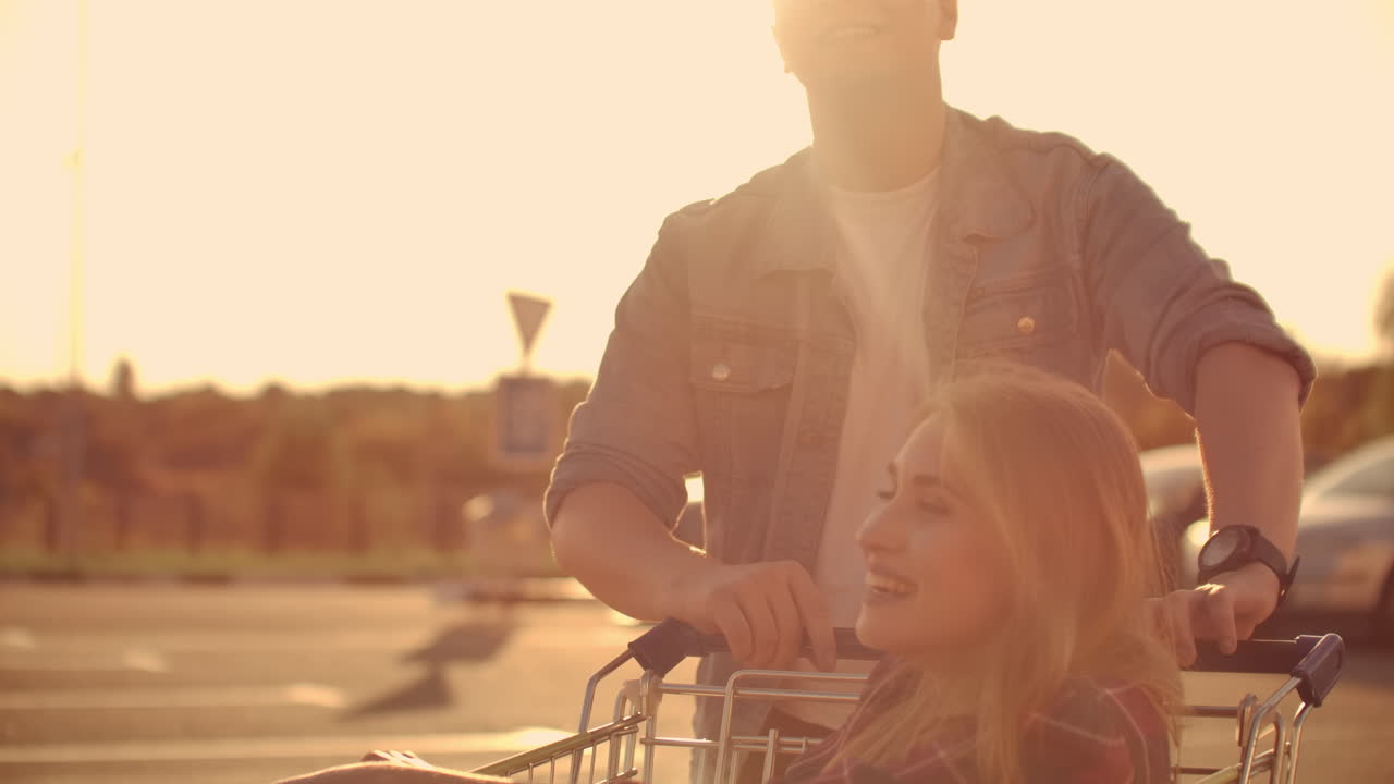 야외 주차장에서 쇼핑 카트와 함께 타고 있는 커플. 슈퍼마켓 근처의 외부 주차장에 쇼핑 카트를 타고 재미있는 젊은 스타일리시 쿠페