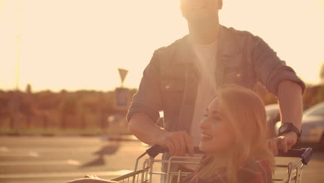 couple riding with shopping cart on the parking outdoors. young stylish coupe having fun riding with shopping cart on the outdoor parking near the supermarket