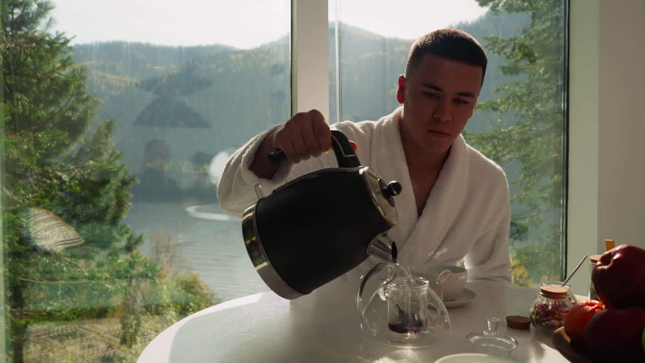 한 남자가 창문 에 있는 차병에 뜨거운 물을 부어 놓는다. 가정용 옷을 입은 조용한 남자가 차가운 액체로 물병을 채우고 자연의 장면에 맞춰 음료를 는다.