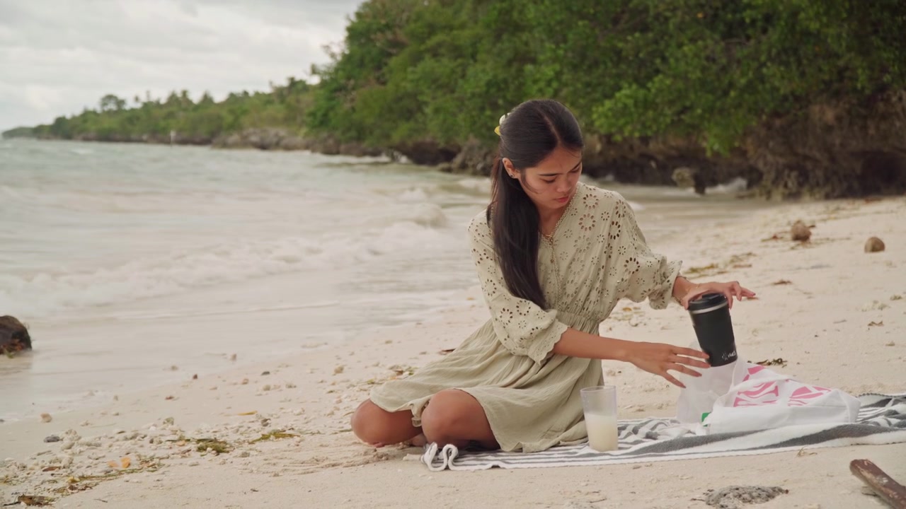 Donna asiatica in spiaggia che prepara un picnic su un telo vicino alla battigia, cielo coperto e onde leggere, stendendo una coperta, posizionando una tazza e degli spuntini, vestito di lino casual con capelli intrecciati, fogliame tropicale.