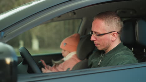 a man in a green jacket sits in the driver's seat of a car, gazing intently out the window with a focused expression. his daughter, dressed in a pink beanie and jacket, is absorbed in her smartphone